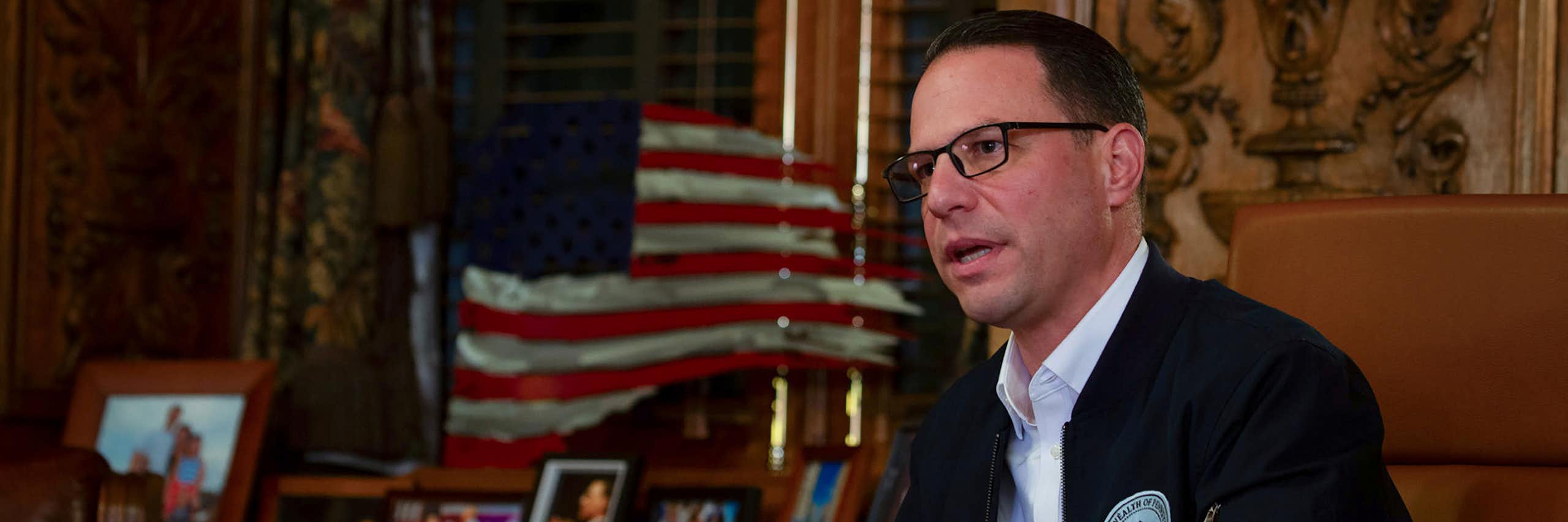 Man in suit jacket and white shirt and wearing glasses sits at table with American flag in background