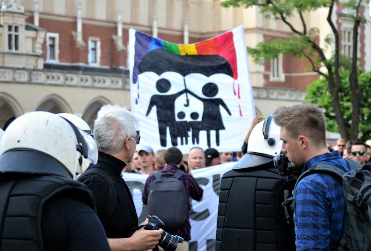 Crowd of people, center of which is a sign depicting silhouette figures of a man and a woman holding an umbrella shielding two children from a rainbow