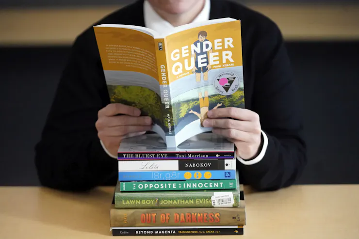 Person reading the book 'Genderqueer' atop a stack of other challenged books