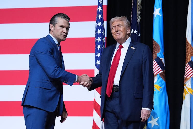 A younger man in a blue suit shakes hands with an older man in a blue suit and red tie who smiles smugly.