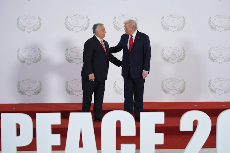 A short stout man in a suit shakes hands with a taller rotund man with whiteish-blond hair.