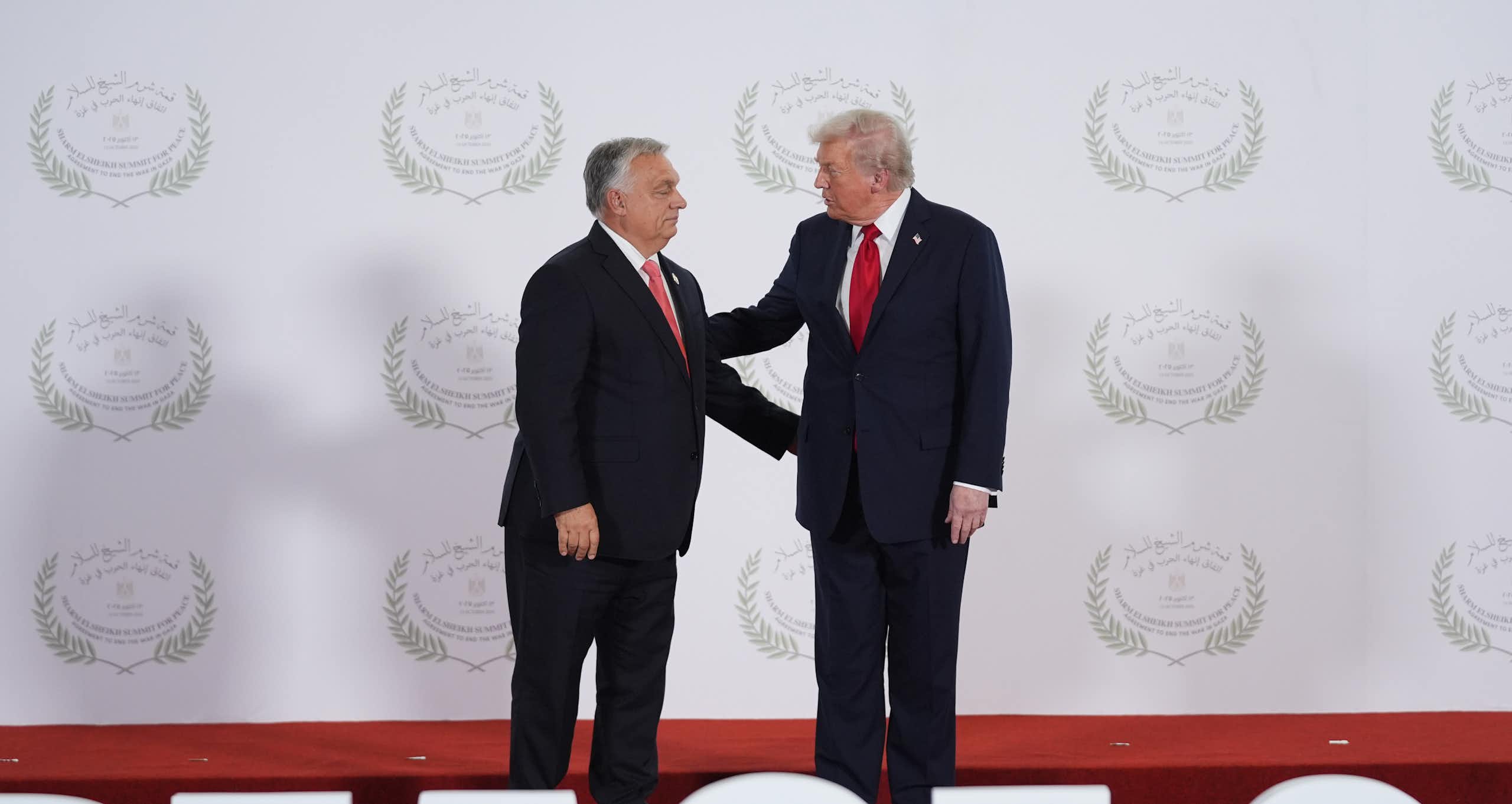 A short stout man in a suit shakes hands with a taller rotund man with whiteish-blond hair.