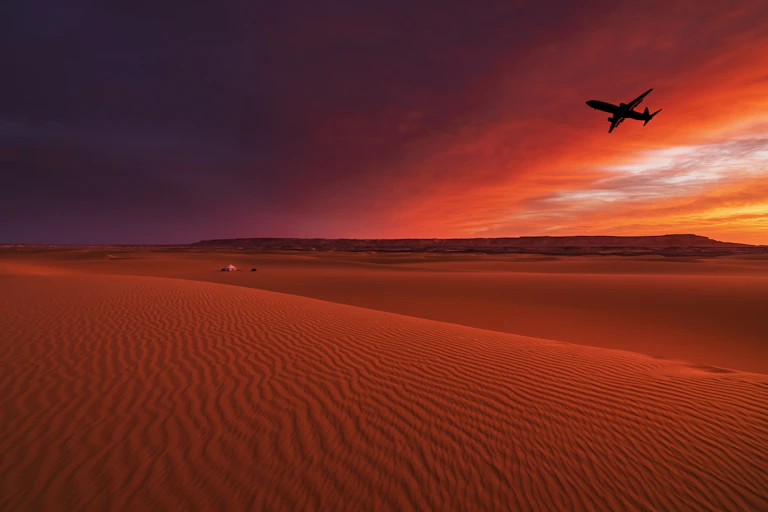 A large plane flies over a desert in the sunset with gold and pink sky in the background