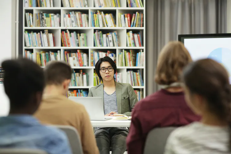 A teacher sits at the front of a high school class with a laptop on their desk. Shelves of books are behind the teacher.