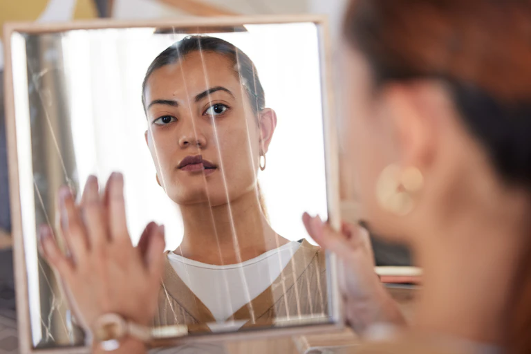 A young woman looking somberly into a broken mirror