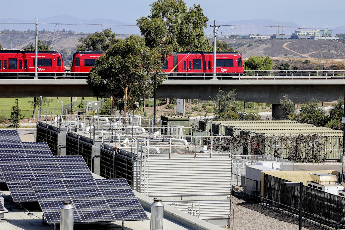 A group of solar panels sits next to several large metal containers, as a train rolls past in the background.