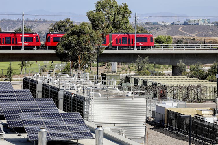 A group of solar panels sits next to several large metal containers, as a train rolls past in the background.