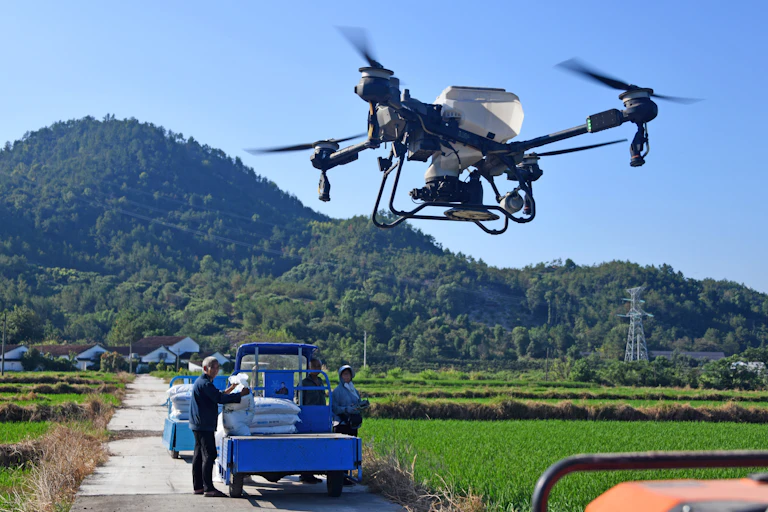 People stand next to a small truck while a four-propeller drone with a white tank to hold liquid flies overhead.