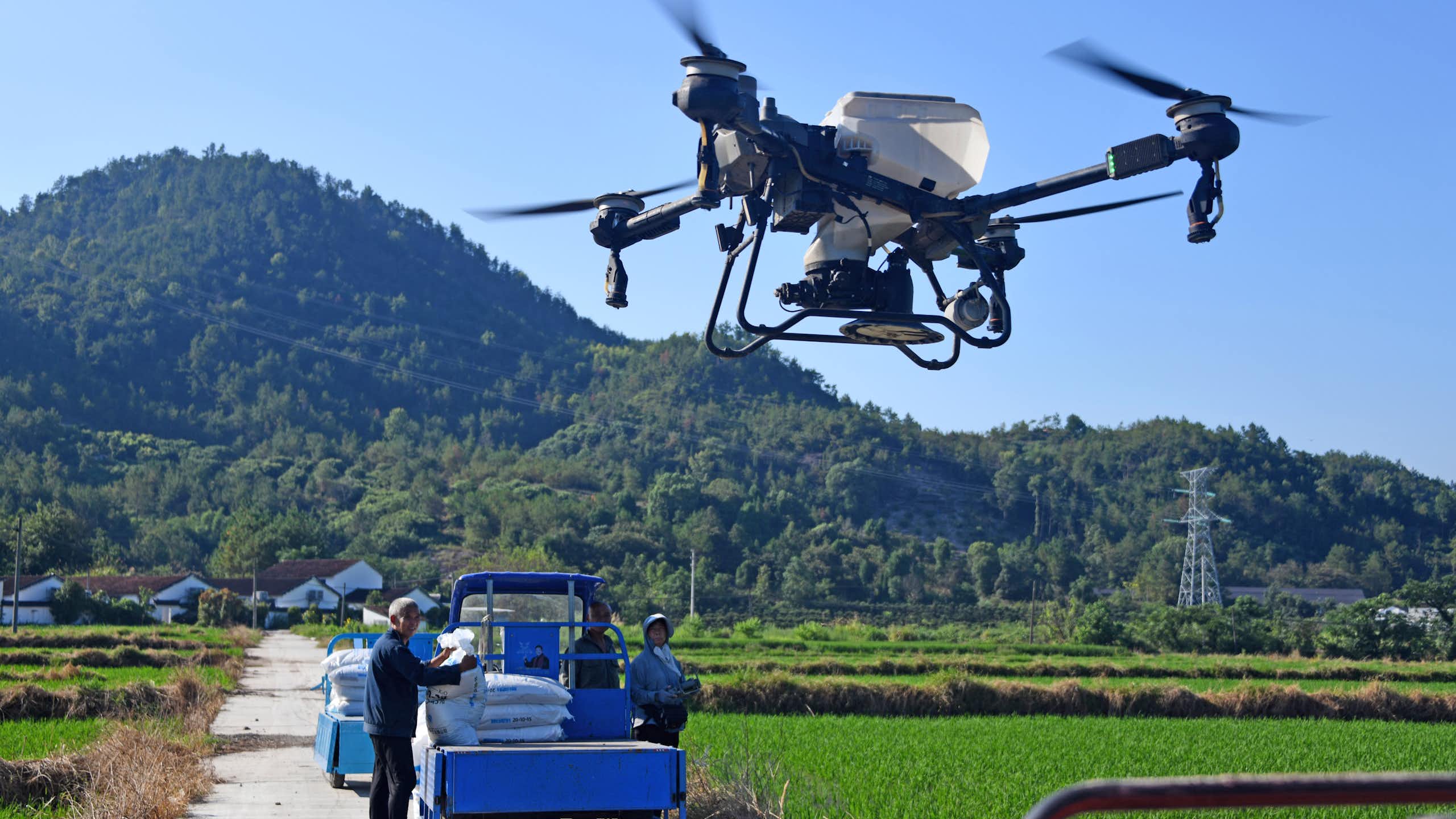 People stand next to a small truck while a four-propeller drone with a white tank to hold liquid flies overhead.