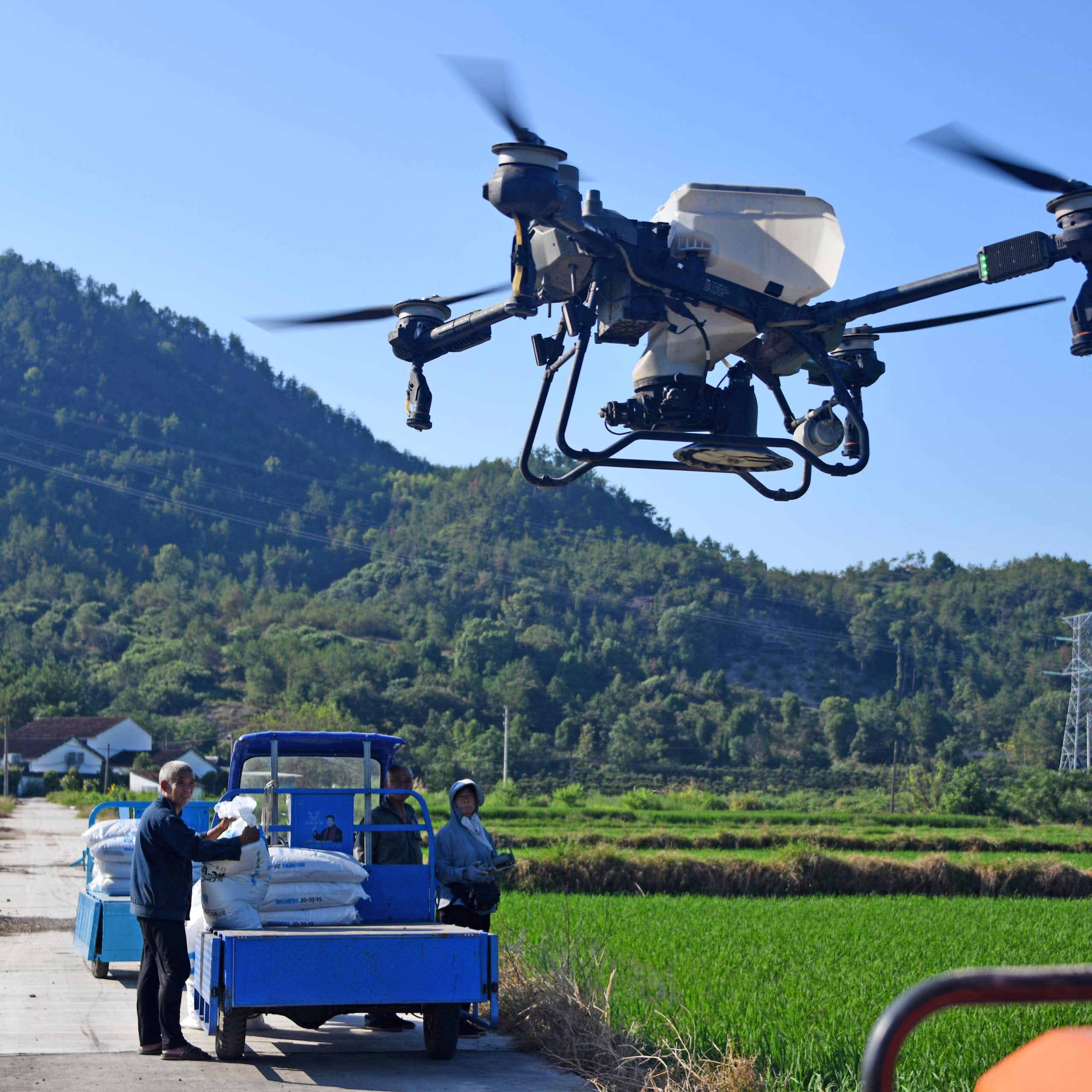 People stand next to a small truck while a four-propeller drone with a white tank to hold liquid flies overhead.