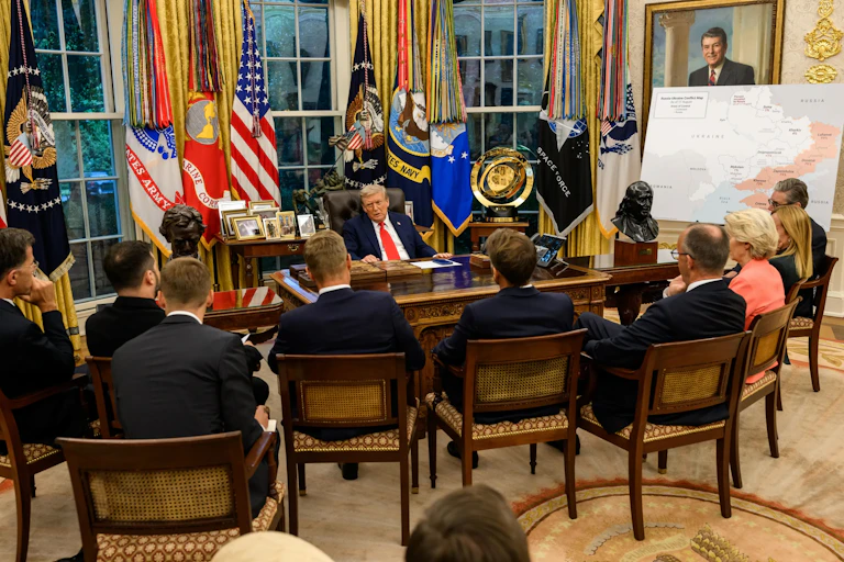 A man sits at a desk talking to several other people. A map is behind him.