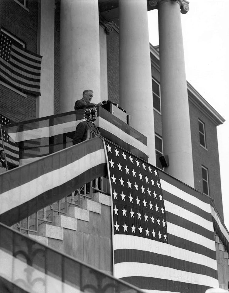In a black-and-white photograph, President Franklin D. Roosevelt stands on the steps of a new building, draped with American flags.