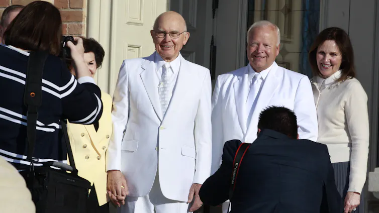 Un homme vêtu d’un costume blanc et d’une cravate se tient debout devant le porche d’un temple. Il est pris en photo par une femme qui porte un pull à rayures noires et blanches