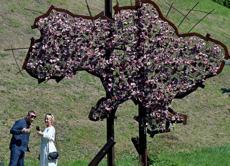 Two people take a selfie in front of an outline of a map made with flowers.