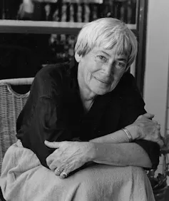 An older woman with a smiling intelligent face next to a bookcase.