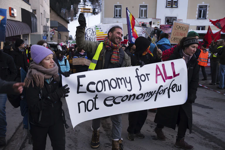 A young man and two women in winter clothing hold a sign that says, 'Economy for ALL not economy for few.'