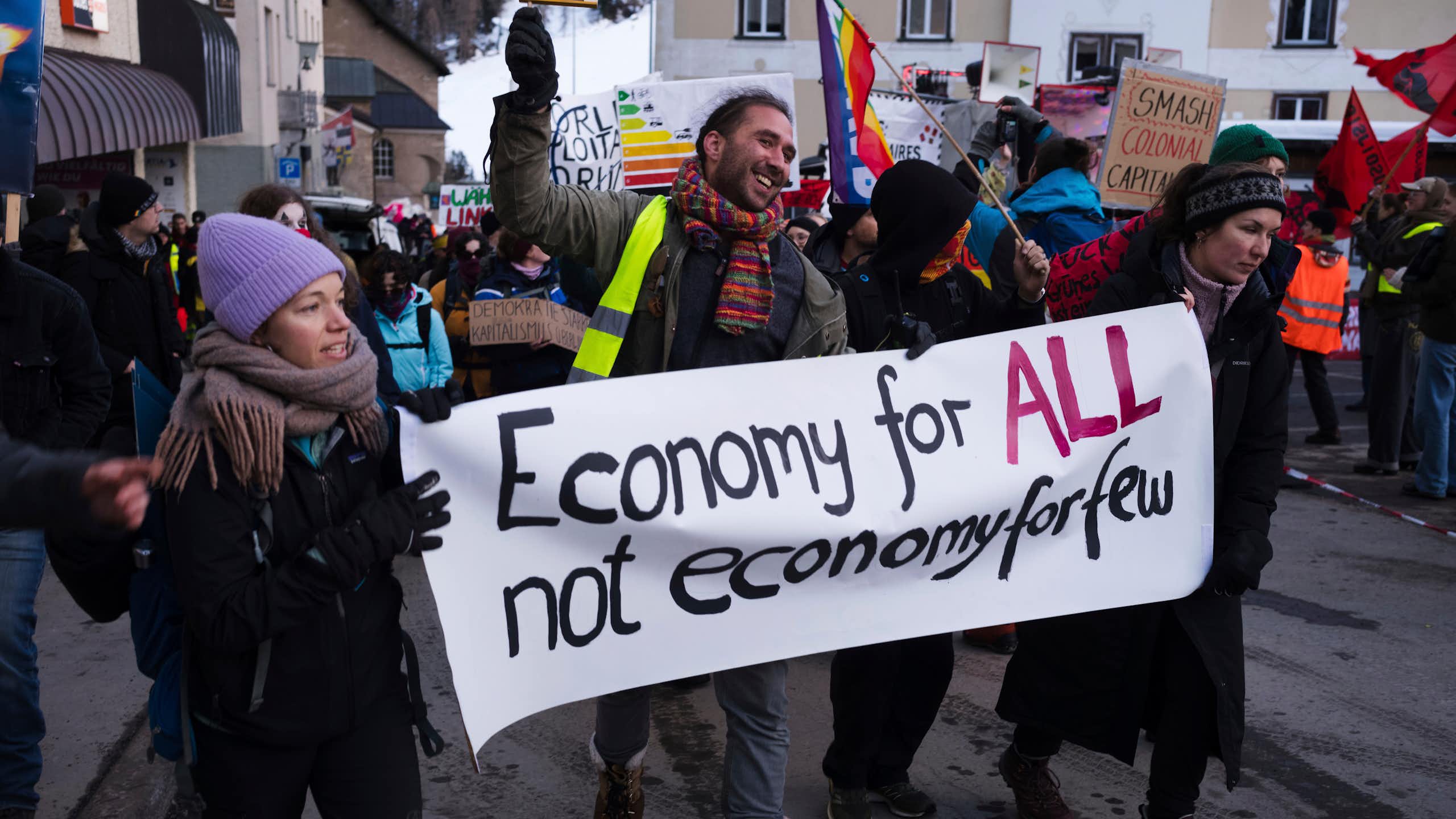 A young man and two women in winter clothing hold a sign that says, 'Economy for ALL not economy for few.'