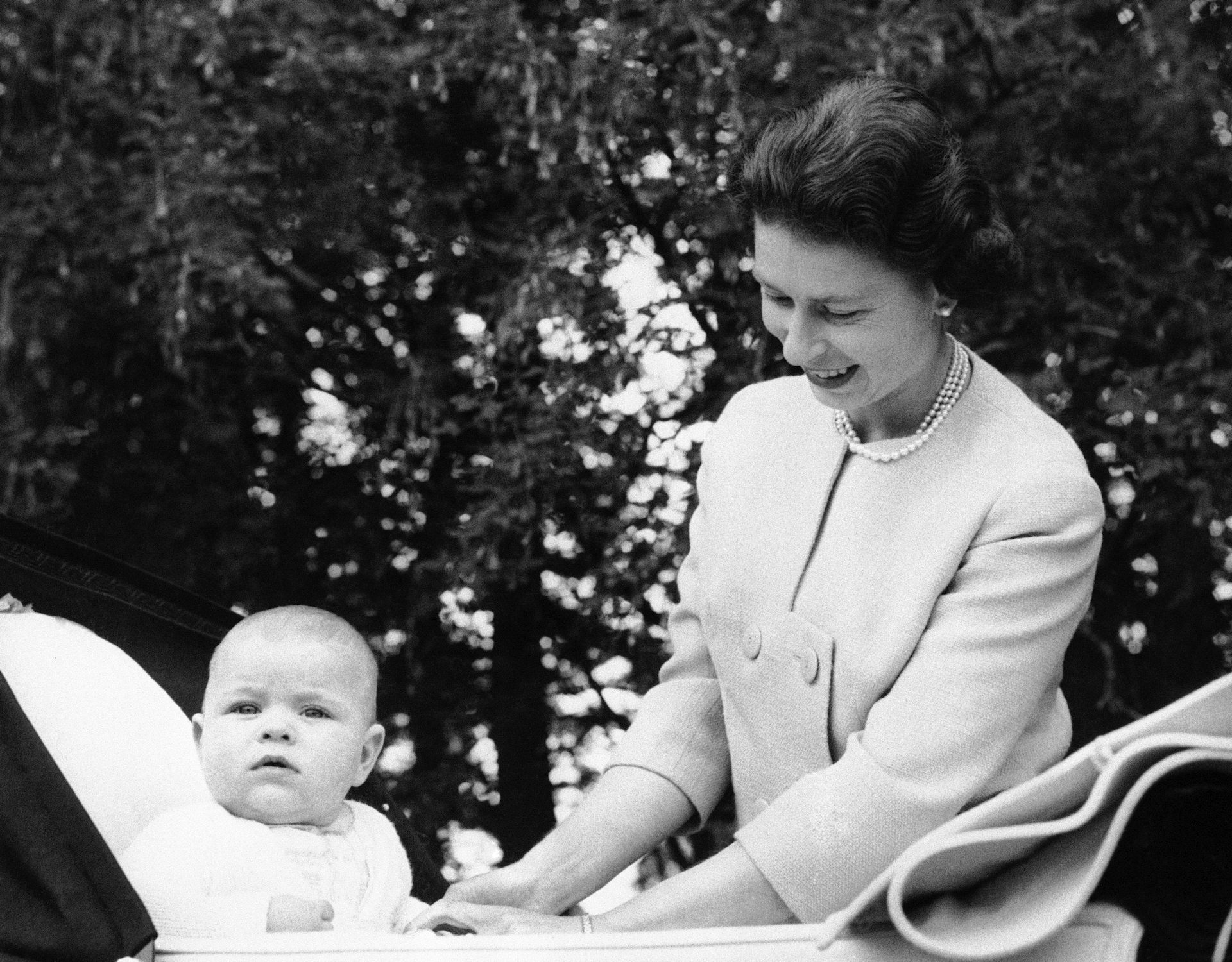 A black-and-white photo shows a round-faced baby sitting in his carriage as his mother smiles down at him.