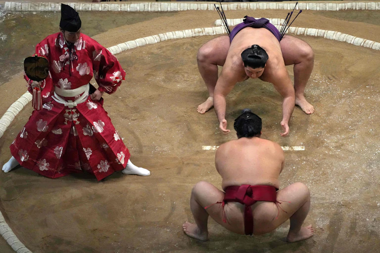 Two men in silk loincloths crouch on a dirt floor as another man in ornate red robes looks on.
