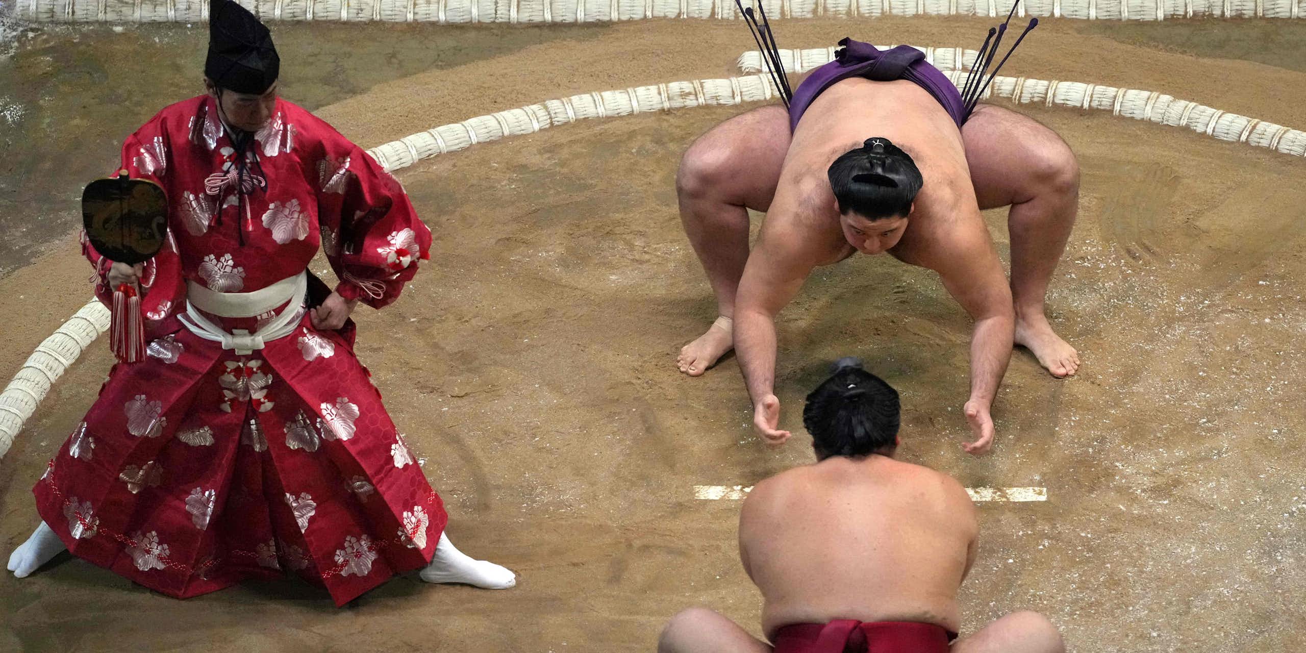 Two men in silk loincloths crouch on a dirt floor as another man in ornate red robes looks on.