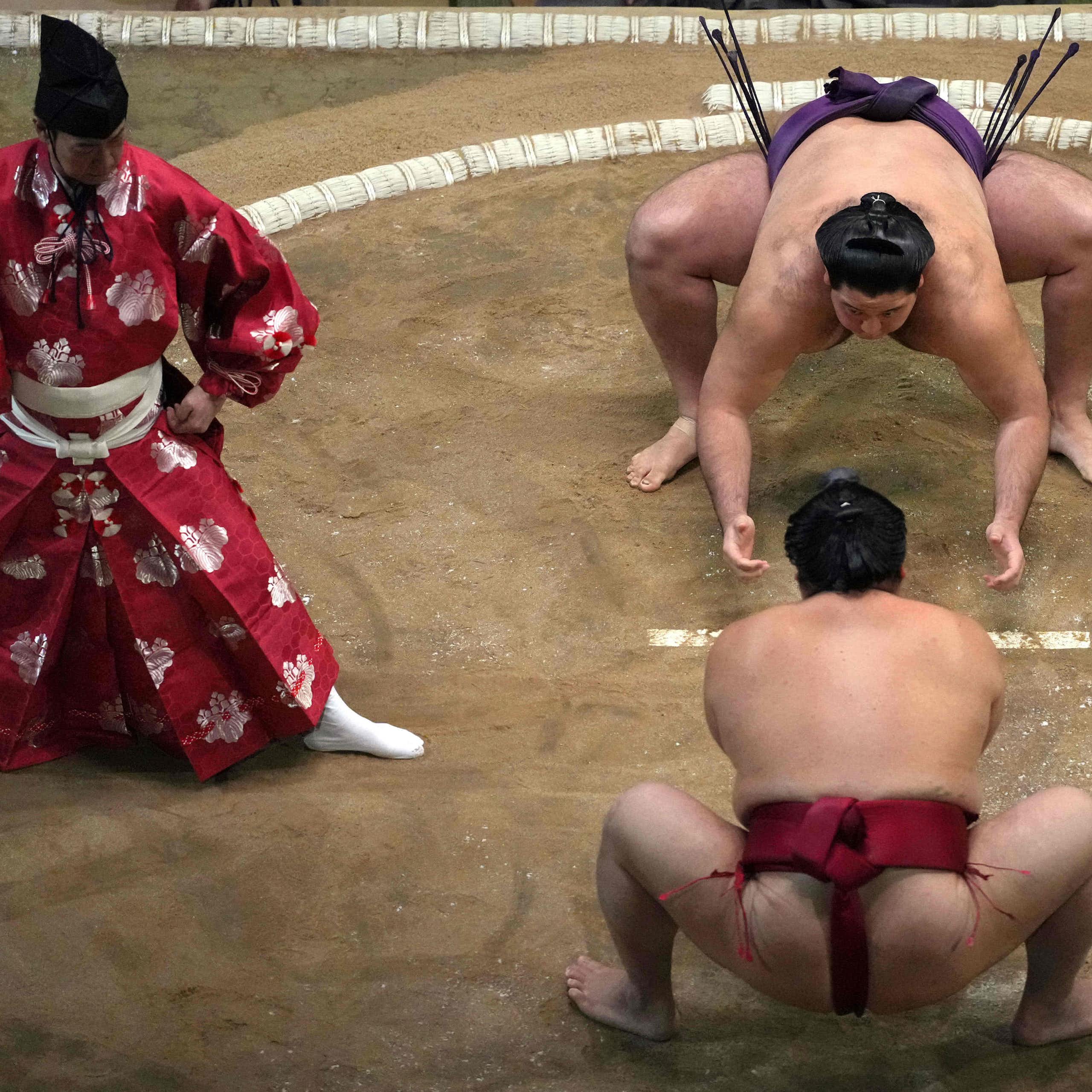 Two men in silk loincloths crouch on a dirt floor as another man in ornate red robes looks on.