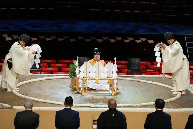 Two men in white robes bow, standing on the side of a dirt ring, as another man in white robes sits between them.