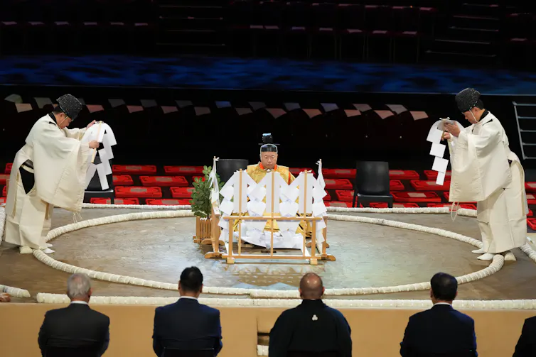 Two men in white robes bow, standing on the side of a dirt ring, as another man in white robes sits between them.