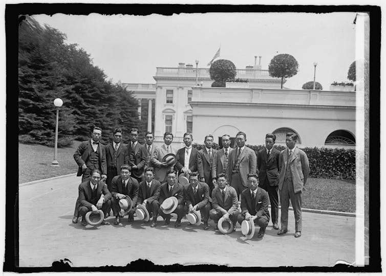 A black and white photo shows two rows of men in suits posing outside a large white building.