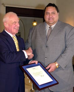 A man in a blue suit shakes hands with a much larger man in a gray suit.