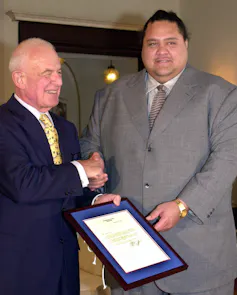 A man in a blue suit shakes hands with a much larger man in a gray suit.