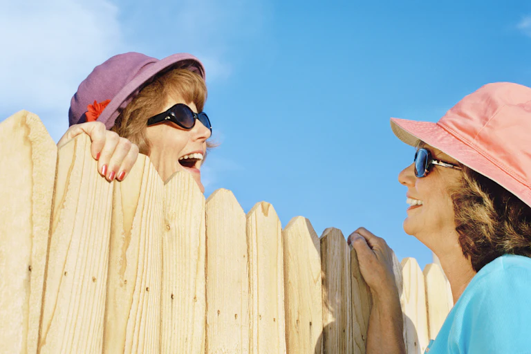 Two ature women in hats talk over a fence.