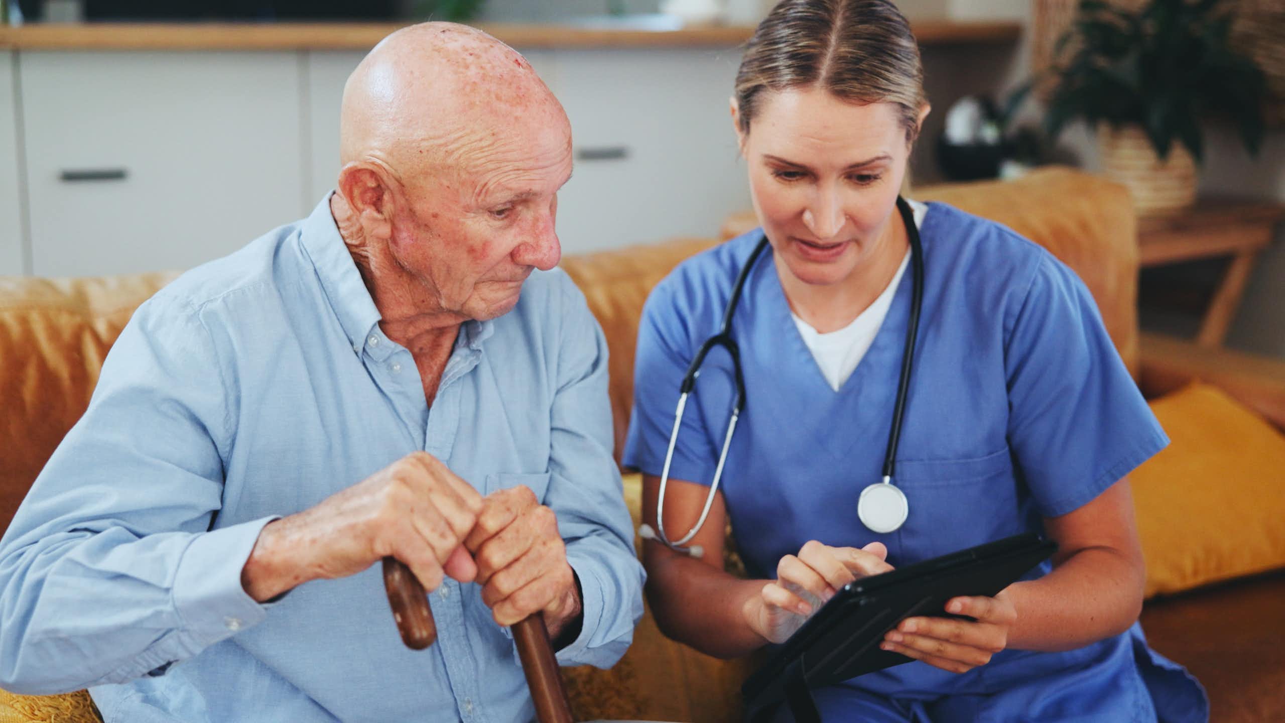 Une soignante est assise à côté d’un monsieur âgé pour des soins délivrés à son domicile