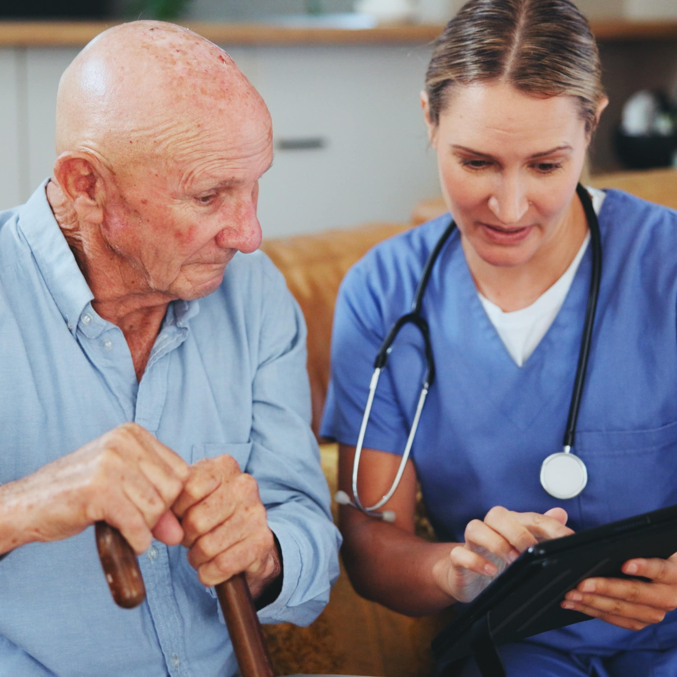 Une soignante est assise à côté d’un monsieur âgé pour des soins délivrés à son domicile