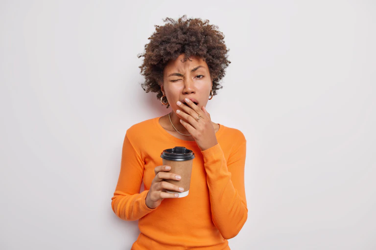 Woman wearing orange top yawning and holding takeaway coffee cup.