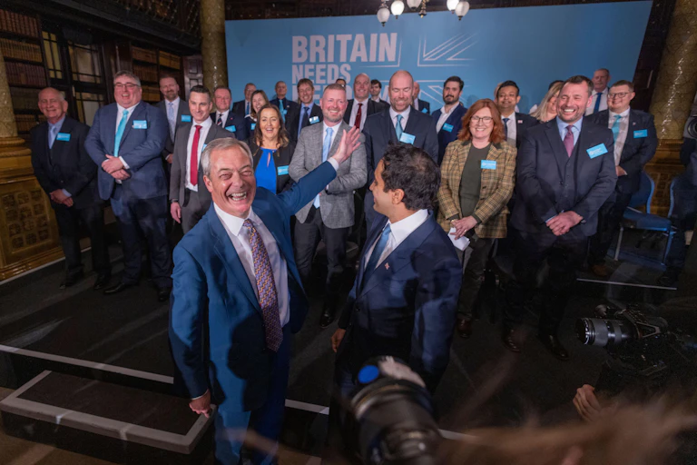 Nigel Farage and Zia Yusuf gesturing towards a stage full of councillors.