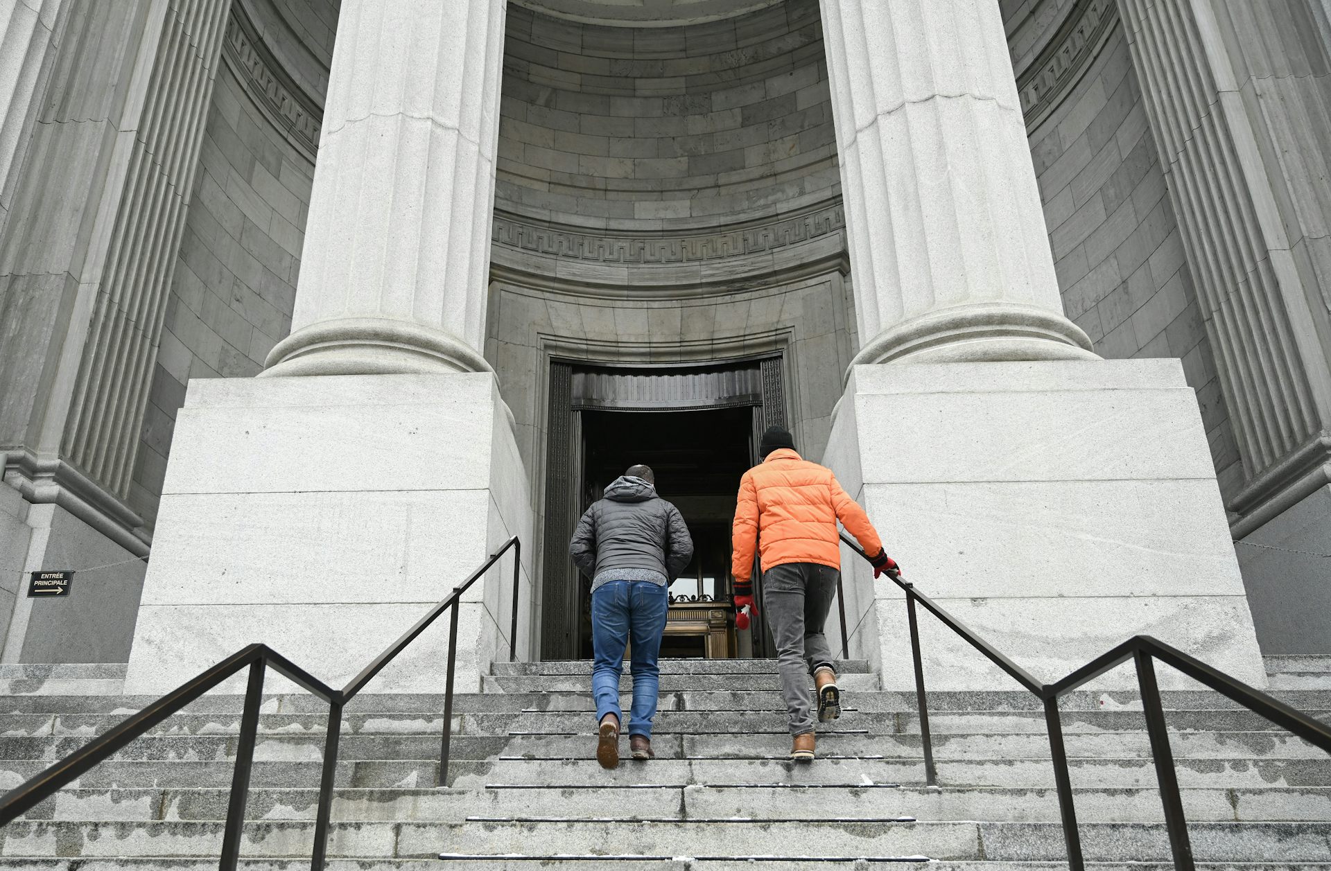 Two people walk up the steps of a courthouse with large stone pillars on either side of a stone staircase.