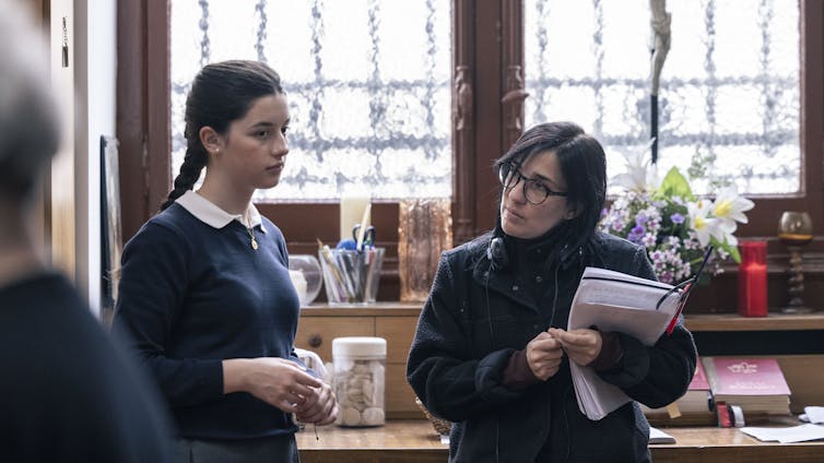 Una chica vestida de uniforme atiende a algo fuera de campo mientras una mujer que sujeta muchos papeles a su lado la mira.