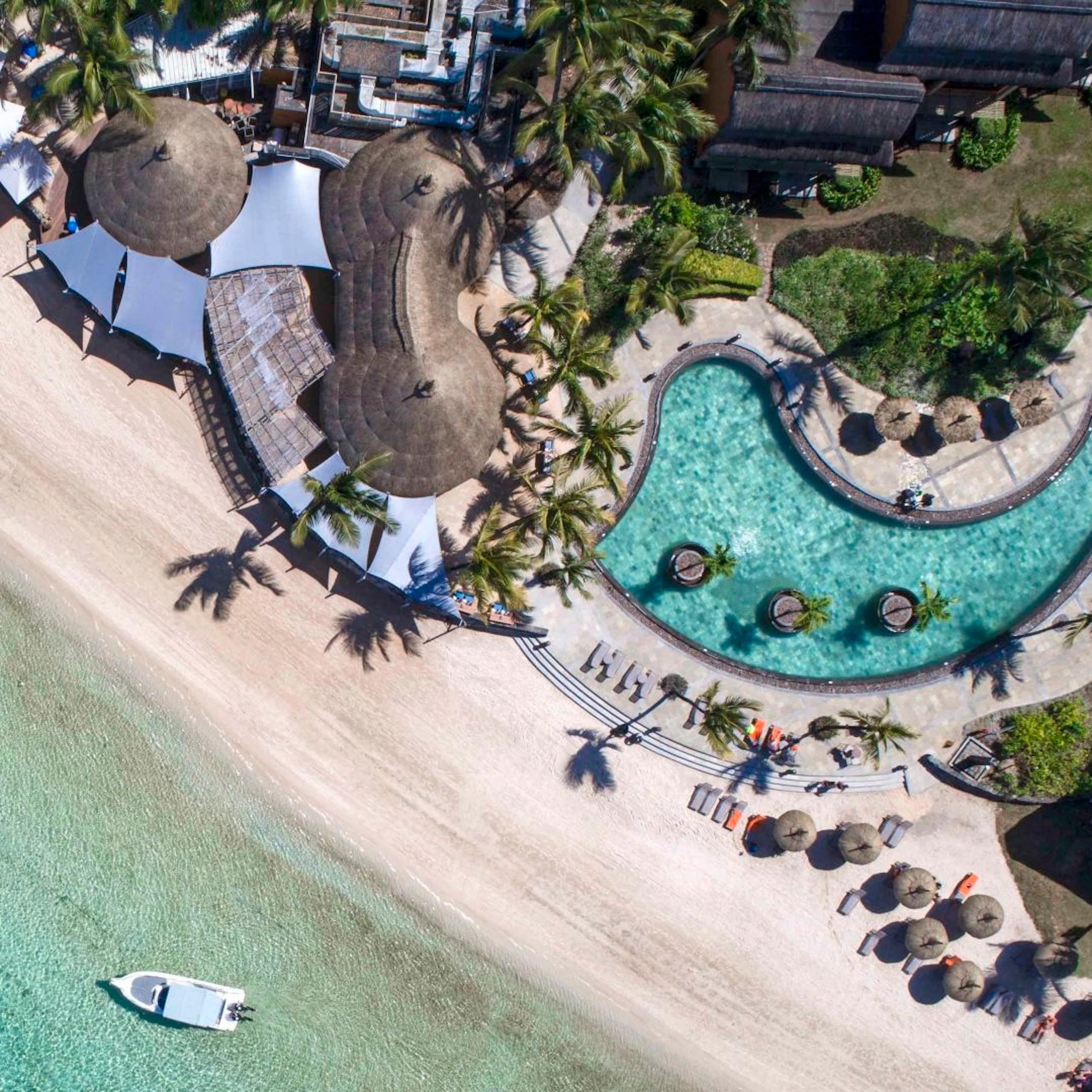 An aerial photo of a curved structure with large curved pool right on a beach and the sea, where a speedboat is seen.