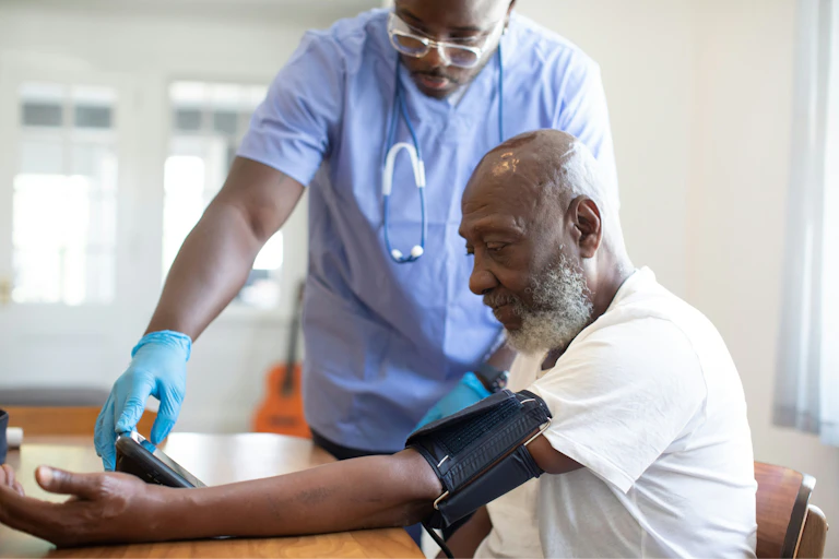 An older patient seated wearing a blood pressure cuff with a younger man taking his blood pressure