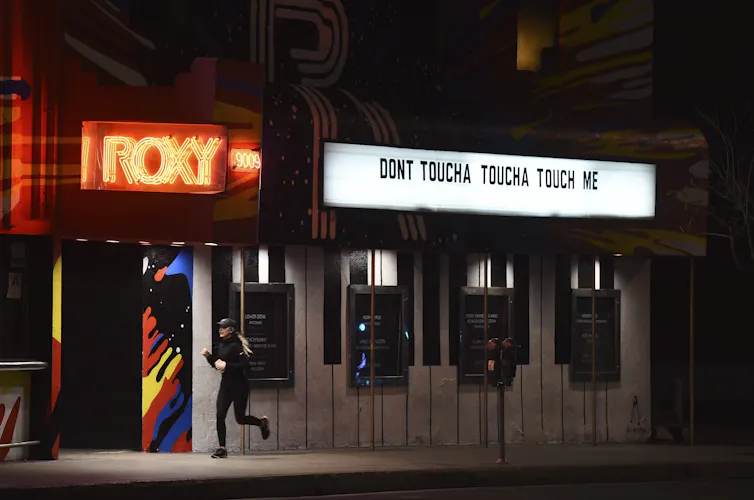 A person jogs past a marquee in the dark that reads 'dontcha dontcha touch me.'