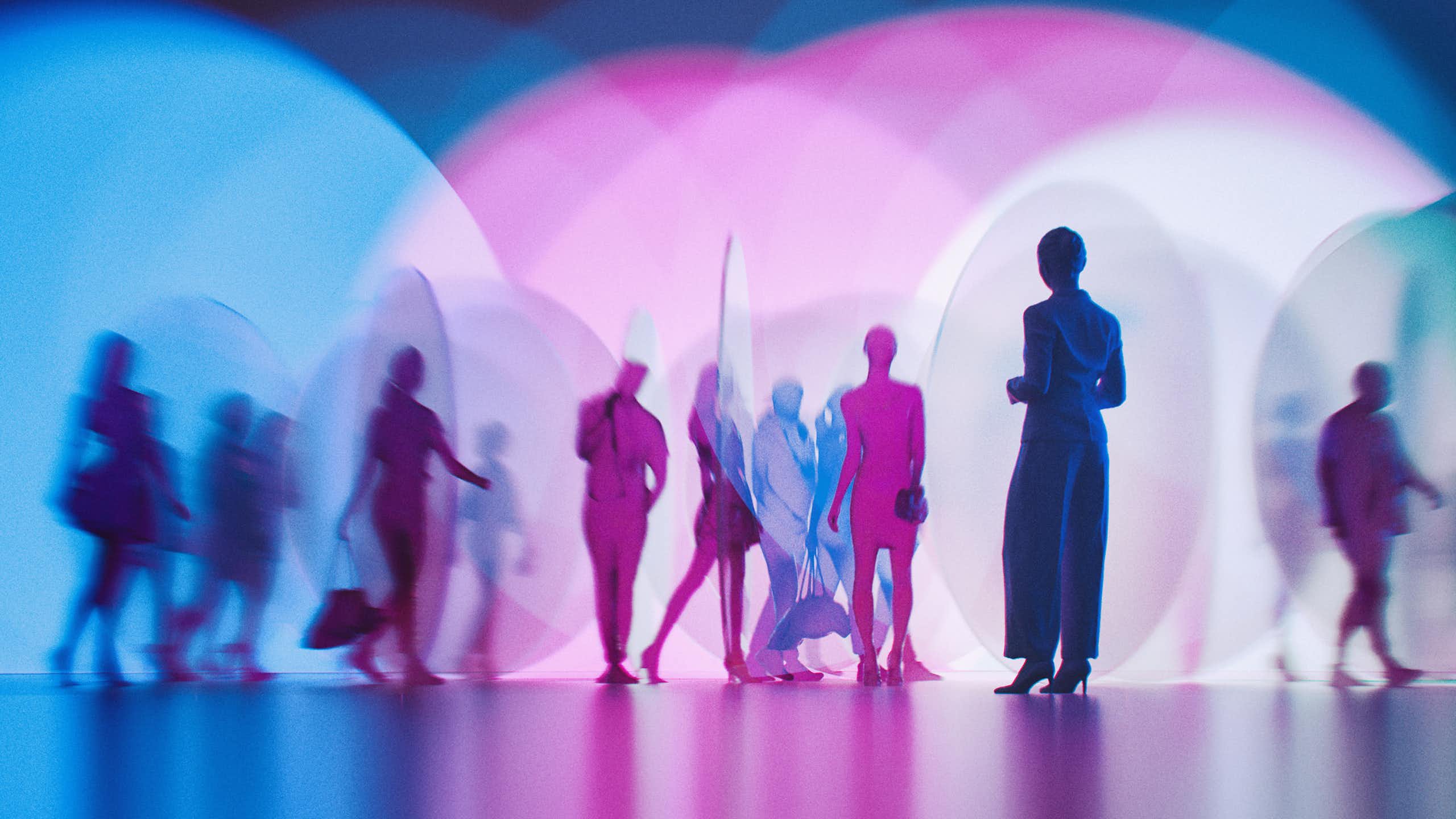 Blurred silhouettes of people walking across a backdrop of overlapping blue and pink lights, one silhouette standing slightly apart in the foreground