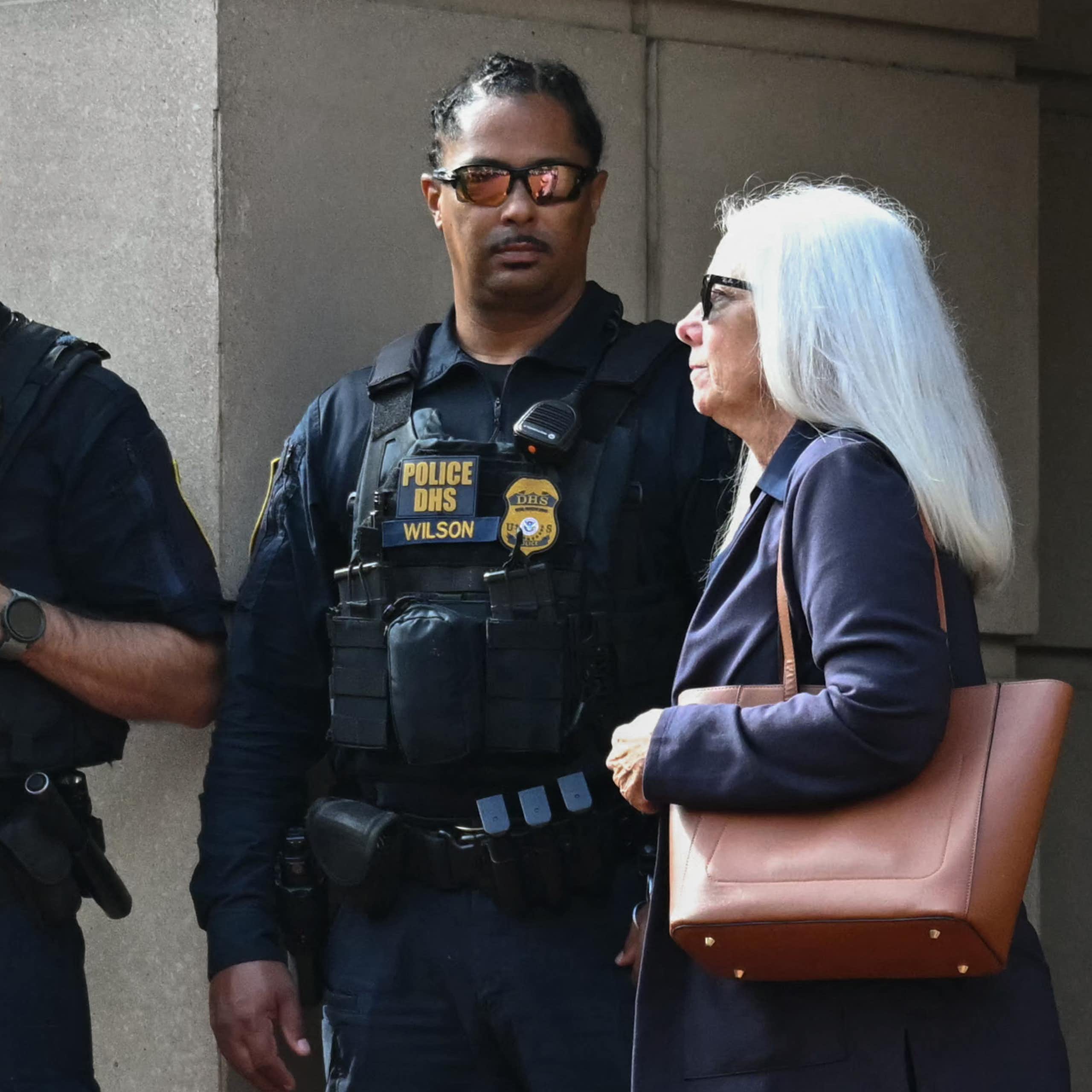 A white-haired woman in a jacket and holding a shoulder bag walks past two law enforcement officers.