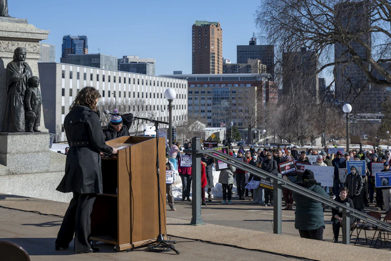 A speaker standing at a lectern on a college campus looks across a crowd of people holding signs.
