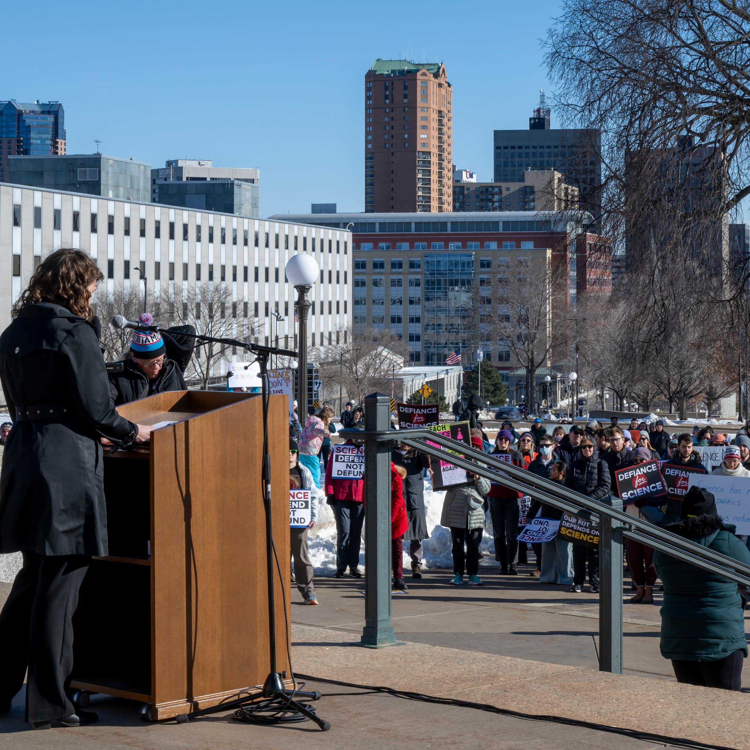 A speaker standing at a lectern on a college campus looks across a crowd of people holding signs.