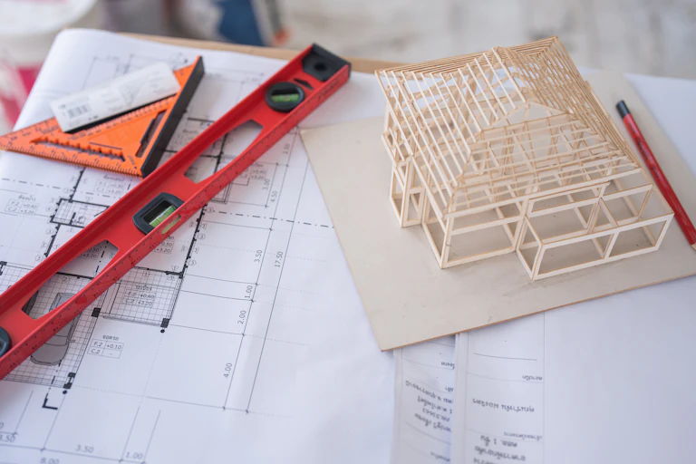 Red and orange tools sit on top of a blueprint, next to a small wooden model of a house.