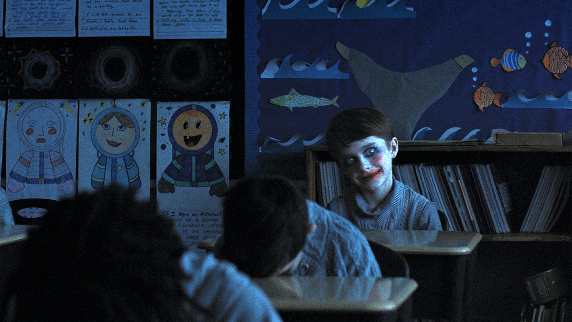 Un niño con la sonrisa pintada mira a la cámara mientras sus compañeros duermen en sus pupitres.