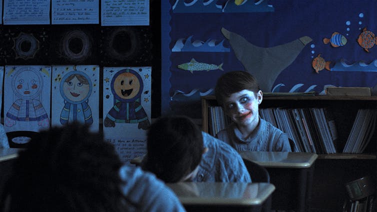 De vampiros a vecinos molestos: cómo ha cambiado la forma de asustarnos en el cine 2 Un niño con la sonrisa pintada mira a la cámara mientras sus compañeros duermen en sus pupitres.