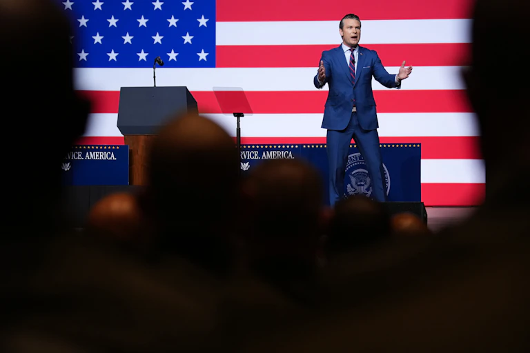 A man on stage, with an American flag behind him, speaks to crowd.