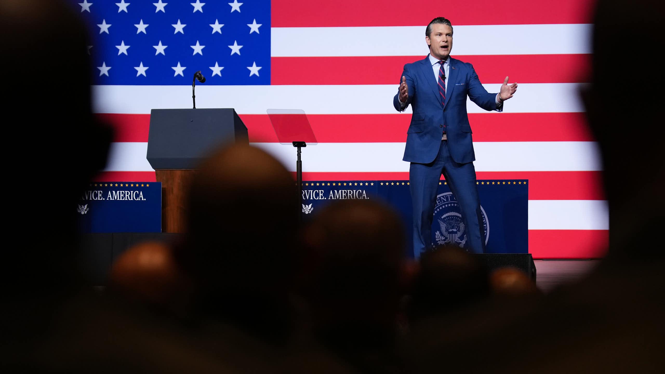 A man on stage, with an American flag behind him, speaks to crowd.