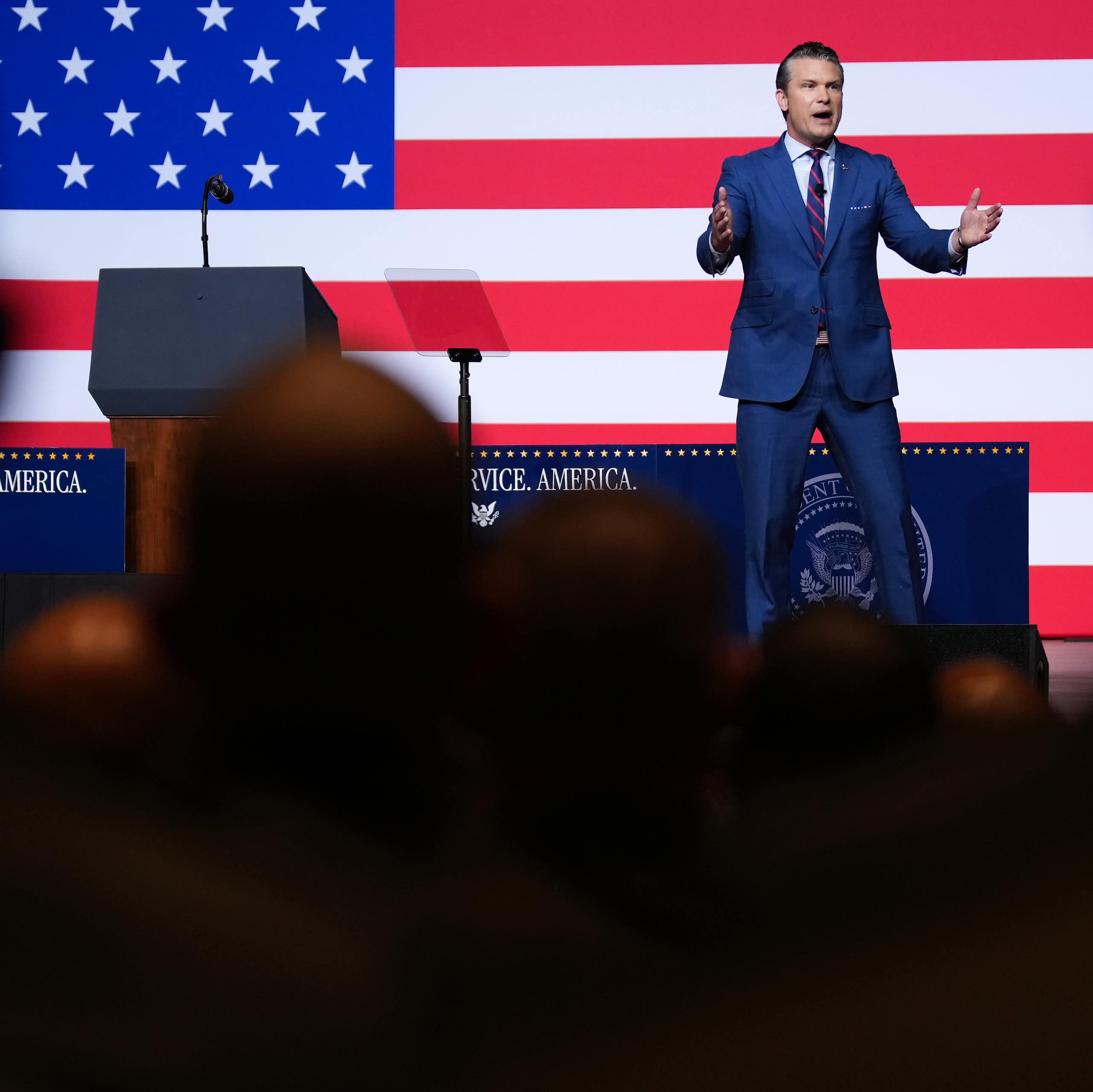 A man on stage, with an American flag behind him, speaks to crowd.